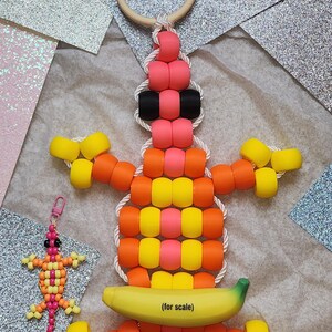 May include: A large, colourful beaded lizard toy with a wooden ring and a smaller beaded lizard keyring. The toy is made of pink, orange, yellow, and black beads. A banana is included for scale, with the text "(for scale)" on it.