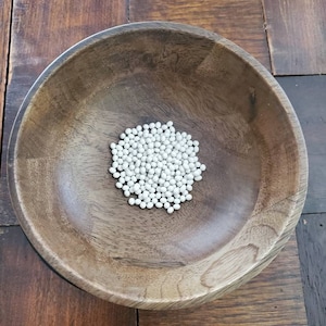 May include: A wooden bowl filled with white round beads. The bowl is on a wooden surface.