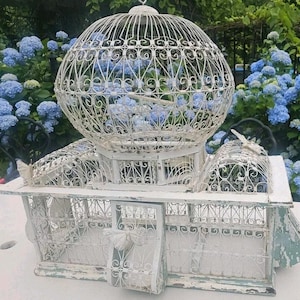Peut inclure: Une cage à oiseaux en métal blanc dotée d'une grande partie supérieure sphérique avec un travail de volutes en fil de fer complexe et une base rectangulaire avec une porte à charnière. La structure présente une finition peinture usée.