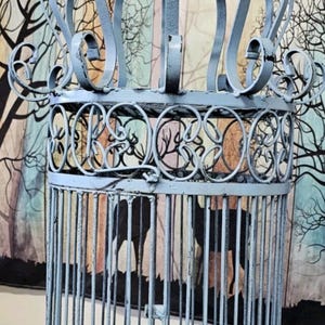 May include: A light blue metal birdcage featuring a cylindrical body with vertical bars and decorative scrollwork patterns. The top is crowned with an arched, open-frame dome structure. The entire piece has a distressed finish and small feet.