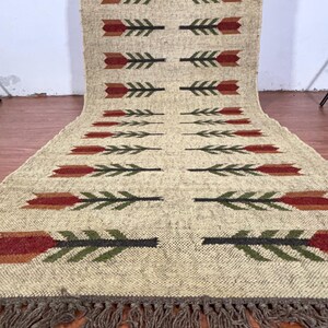 May include: Beige woven runner rug with a repeating pattern of arrows in red, green, and black. The rug has a fringe along the bottom edge. The rug is made of a thick, textured material.