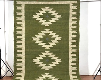 May include: A rectangular green rug with a geometric pattern in cream and gray. The rug has a border and fringe. The geometric design features diamond shapes. The rug is hanging on a black metal frame.