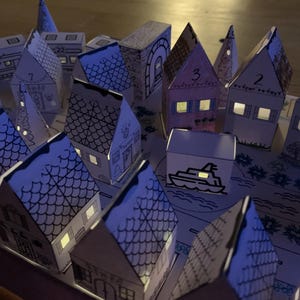 K&ouml;nnte beinhalten: Ein Miniatur-Papierdorf mit beleuchteten Fenstern, das einen warmen Schein erzeugt. Die H&auml;user sind nummeriert und mit komplizierten Details verziert, darunter eine Bootsillustration. Das Gesamtthema ist festlich und charmant, ideal f&uuml;r die Weihnachtsdekoration.