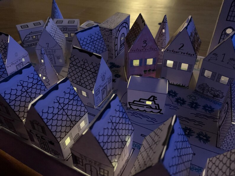 K&ouml;nnte beinhalten: Ein Miniatur-Papierdorf mit beleuchteten Fenstern, das einen warmen Schein erzeugt. Die H&auml;user sind nummeriert und mit komplizierten Details verziert, darunter eine Bootsillustration. Das Gesamtthema ist festlich und charmant, ideal f&uuml;r die Weihnachtsdekoration.