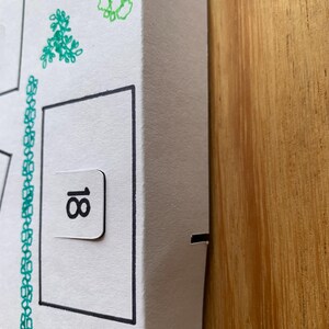 K&ouml;nnte beinhalten: Wei&szlig;er Pappkarton mit schwarzem Rand und der Zahl 18 auf einer kleinen Lasche. Die Box ist mit gr&uuml;nen floralen Illustrationen verziert und steht auf einer Holzoberfl&auml;che.