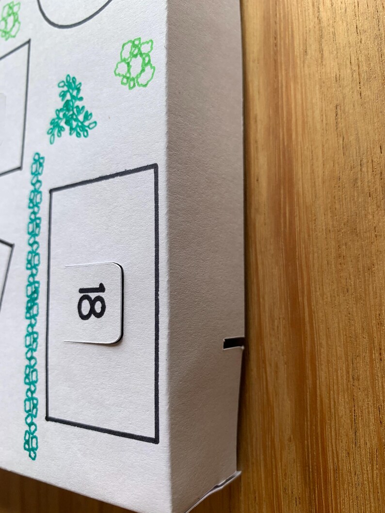 K&ouml;nnte beinhalten: Wei&szlig;er Pappkarton mit schwarzem Rand und der Zahl 18 auf einer kleinen Lasche. Die Box ist mit gr&uuml;nen floralen Illustrationen verziert und steht auf einer Holzoberfl&auml;che.