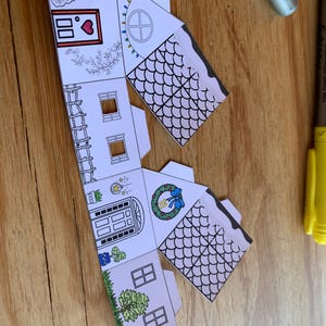 K&ouml;nnte beinhalten: Ein teilweise zusammengebautes Papierhaus mit detaillierten Illustrationen. Das Haus hat eine Eingangst&uuml;r, Fenster und ein Dach mit einem schuppenartigen Muster. Dekorationen umfassen einen Kranz, ein Herz und Lichterketten.