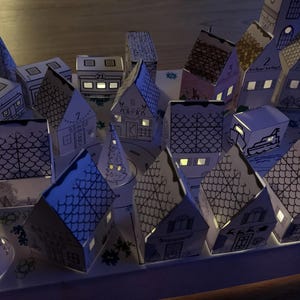 K&ouml;nnte beinhalten: Ein beleuchteter Adventskalender mit einer Pop-up-Dorfszene. Die H&auml;user und anderen Strukturen sind wei&szlig; mit schwarzen Umrissen, und einige haben Nummern. Die Fenster leuchten mit warmem Licht und schaffen eine festliche Atmosph&auml;re. Das Gesamtthema ist Weihnachten.