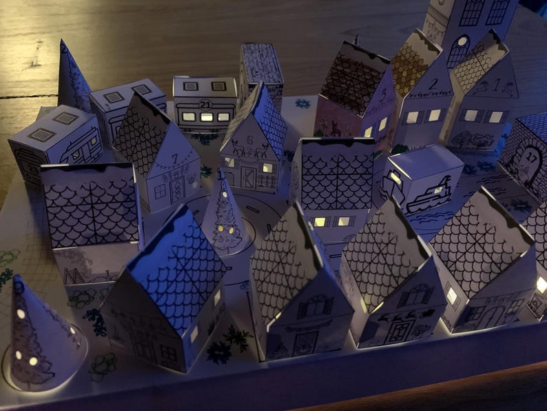 K&ouml;nnte beinhalten: Ein beleuchteter Adventskalender mit einer Pop-up-Dorfszene. Die H&auml;user und anderen Strukturen sind wei&szlig; mit schwarzen Umrissen, und einige haben Nummern. Die Fenster leuchten mit warmem Licht und schaffen eine festliche Atmosph&auml;re. Das Gesamtthema ist Weihnachten.