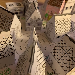 K&ouml;nnte beinhalten: Eine Sammlung kleiner Papierh&auml;user mit Schwarz-Wei&szlig;-Strichzeichnungen, die wie eine Stadt angeordnet sind. Einige H&auml;user haben Nummern und dekorative Details. Die D&auml;cher sind mit einem schuppenartigen Muster versehen, und die gesamte Szene befindet sich auf einer Papierbasis mit einer Stra&szlig;e.