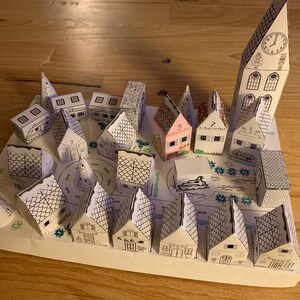 K&ouml;nnte beinhalten: Ein handgefertigtes Papiermodell einer Stadt mit vielen kleinen H&auml;usern, einem Kirchturm mit Uhr und einer zentralen Stra&szlig;e. Die H&auml;user sind wei&szlig; mit schwarzen Umrissen, einige mit rosa oder grauen D&auml;chern. Die Szene ist auf wei&szlig;em Papier gezeichnet.