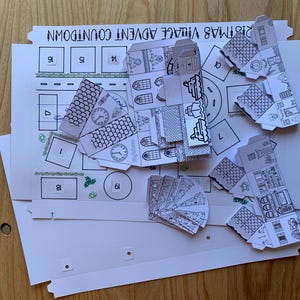 K&ouml;nnte beinhalten: Ein teilweise zusammengebautes Weihnachtsdorf-Adventskalender-Kit. Das Kit enth&auml;lt bedruckte Papierkomponenten zum Bau von Miniaturh&auml;usern und einer Dorflandschaft. Die Komponenten sind schwarz-wei&szlig; und liegen auf einem wei&szlig;en Hintergrund mit dem Text "Christmas Village Advent Countdown".