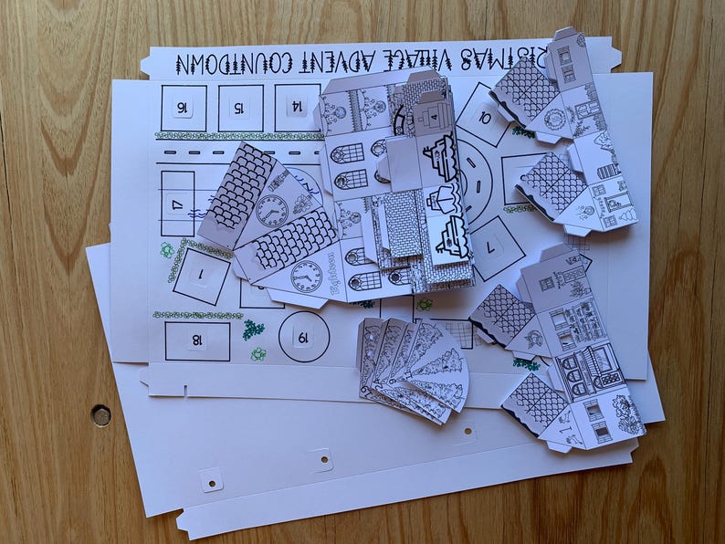 K&ouml;nnte beinhalten: Ein teilweise zusammengebautes Weihnachtsdorf-Adventskalender-Kit. Das Kit enth&auml;lt bedruckte Papierkomponenten zum Bau von Miniaturh&auml;usern und einer Dorflandschaft. Die Komponenten sind schwarz-wei&szlig; und liegen auf einem wei&szlig;en Hintergrund mit dem Text "Christmas Village Advent Countdown".