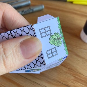 K&ouml;nnte beinhalten: Ein teilweise gebautes Papierhaus mit wei&szlig;er Fassade, zwei quadratischen Fenstern und einer gr&uuml;nen Baumillustration. Das Dach ist hellviolett und die R&auml;nder sind mit gr&uuml;nem Gras verziert. Das Haus wird von einer Hand gehalten.