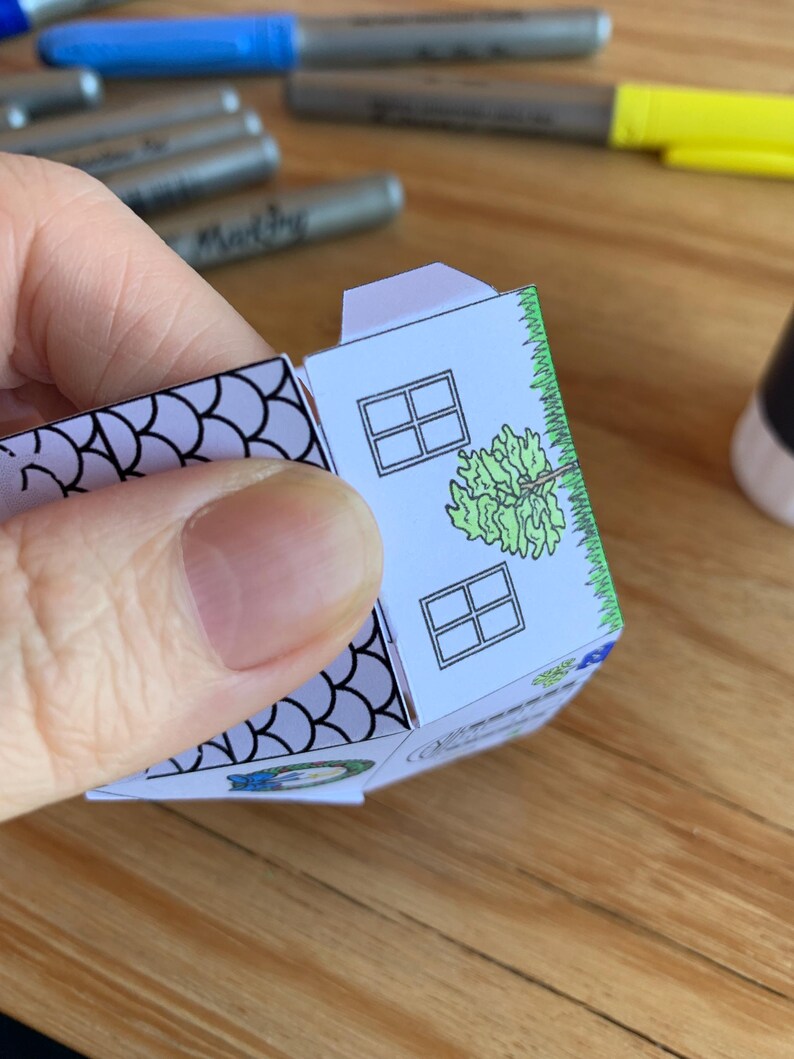 K&ouml;nnte beinhalten: Ein teilweise gebautes Papierhaus mit wei&szlig;er Fassade, zwei quadratischen Fenstern und einer gr&uuml;nen Baumillustration. Das Dach ist hellviolett und die R&auml;nder sind mit gr&uuml;nem Gras verziert. Das Haus wird von einer Hand gehalten.