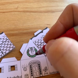 K&ouml;nnte beinhalten: Eine Person bemalt ein Papierhaus mit einem roten Marker. Das Haus hat einen Kranz an der Eingangst&uuml;r und andere Weihnachtsdekorationen. Das Haus ist wei&szlig; mit schwarzen Umrissen, und die Person f&uuml;gt dem Kranz Farbe hinzu.