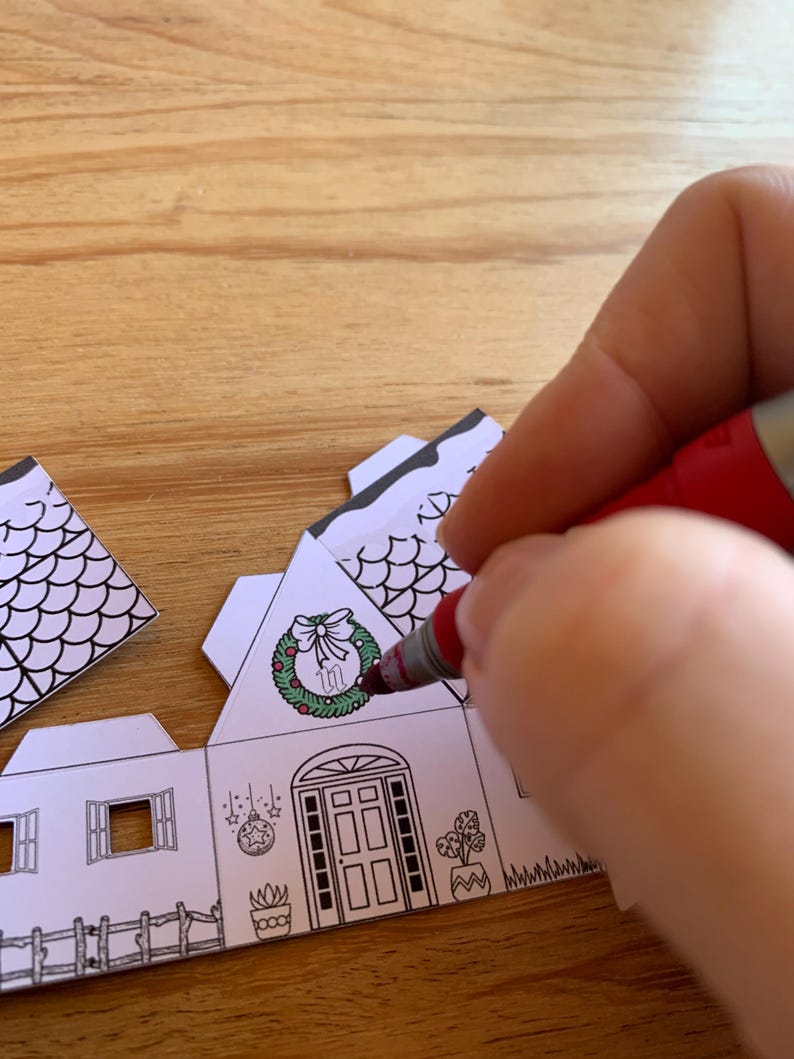 K&ouml;nnte beinhalten: Eine Person bemalt ein Papierhaus mit einem roten Marker. Das Haus hat einen Kranz an der Eingangst&uuml;r und andere Weihnachtsdekorationen. Das Haus ist wei&szlig; mit schwarzen Umrissen, und die Person f&uuml;gt dem Kranz Farbe hinzu.
