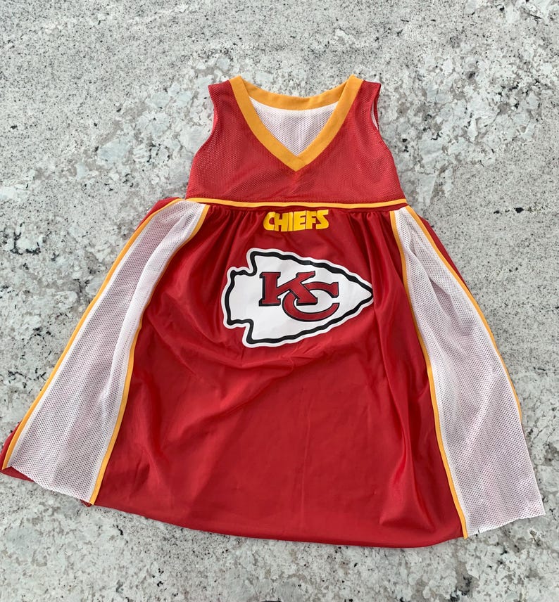 May include: A red and white dress with a Chiefs logo and the word "CHIEFS" in yellow. The dress has a V-neck and white mesh panels with yellow trim. The dress is sleeveless.