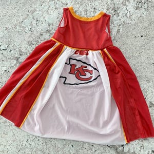 May include: A red and white dress with a Kansas City Chiefs logo. The sleeveless dress has a red mesh overlay with yellow trim and a white panel in the center. The Chiefs logo is in red and black.