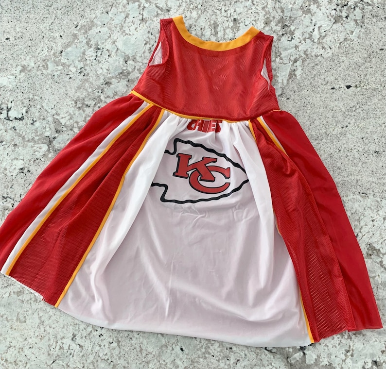 May include: A red and white dress with a Kansas City Chiefs logo. The sleeveless dress has a red mesh overlay with yellow trim and a white panel in the center. The Chiefs logo is in red and black.