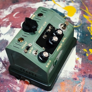 Puede incluir: Un pedal de efectos verde y negro con perillas, interruptores y el texto "Magic Clip" y "Gain". El pedal, de forma rectangular, se utiliza probablemente para la producción o interpretación musical.