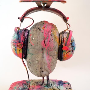 Può includere: Scultura astratta di un volto con cuffie. La scultura è realizzata in cemento e metallo, con spruzzi di vernice colorati in rosa, blu, giallo e arancione. Le cuffie sono dipinte con colori simili.