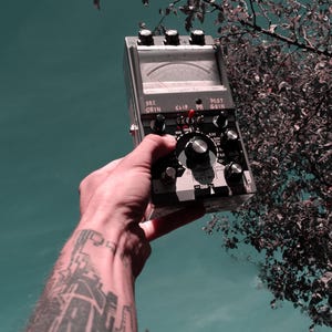 May include: A grey electronic audio signal processor featuring a central analogue needle gauge, three top-mounted control knobs, and a large central dial. The device includes labels such as PRE GRIN, CLIP, and POST GRIN, with a small red toggle switch.