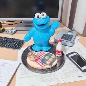 May include: A bright blue Cookie Monster figurine stands on a circular tray next to a small glass bowl of chocolate chip cookie dough with a silver spoon, a metal muffin tin with four cookies, and a miniature white milk bottle with a red cap.
