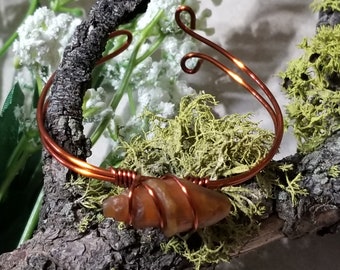 May include: A copper wire bracelet featuring a brown agate stone wrapped in copper wire. The bracelet is shown on a piece of driftwood with moss and yellow flowers.
