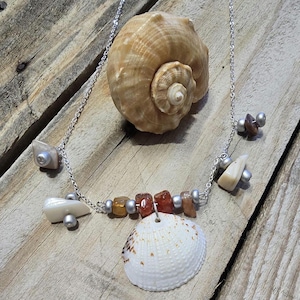 May include: A silver chain necklace with a pendant of small, colorful stones and seashells. The necklace features a variety of shapes and colors, including orange, brown, and white. A large seashell is in the background.