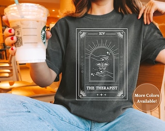 May include: A dark gray t-shirt with a white graphic of a tarot card. The card is labeled "XIV THE THERAPIST" and features a hand with an eye and a butterfly. The text "More Colors Available!" is in a white circle on the bottom right of the shirt.