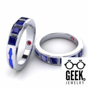 May include: Two silver rings with blue and red accents. Each ring features a row of square blue gemstones and a small red gemstone. The rings have a blue arrow design on the side. The Geek Jewelry logo is in the bottom right corner.