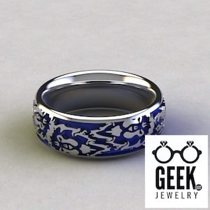 Peut inclure: Une bague argentée avec un intérieur bleu et un motif décoratif autour de l'anneau. La bague a une finition polie et un design moderne. Le logo Geek Jewelry est dans le coin inférieur droit.