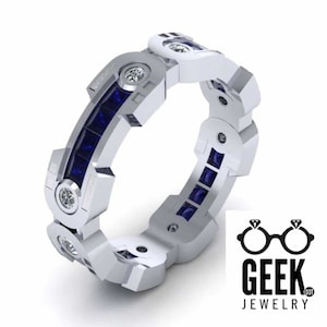 May include: A white metal ring with a geometric design, featuring alternating sections of round diamonds and square, deep blue gemstones. The ring has a modern, industrial aesthetic. The Geek Jewelry logo is in the bottom right corner.