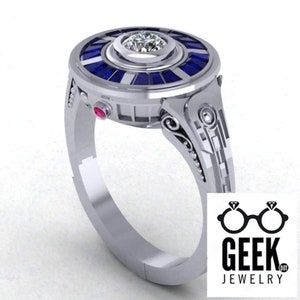May include: A silver ring with a circular design featuring a central clear gemstone surrounded by blue stones. The ring has a detailed band with decorative elements and a small red stone on the side. The Geek Jewelry logo is in the bottom right corner.