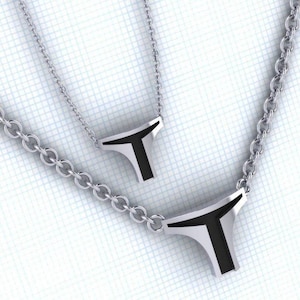 Peut inclure: Deux colliers argentés avec des pendentifs en forme de T géométriques. Les pendentifs ont un insert en émail noir et sont suspendus à de fines chaînes. Les colliers sont présentés sur un fond quadrillé blanc.