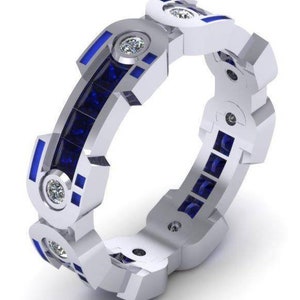 May include: A silver-colored ring with a geometric design. The ring features alternating sections of round, clear gemstones and rectangular, dark blue gemstones. The ring's design includes blue accents and is set against a white background.