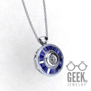May include: A silver pendant necklace with a circular design. The pendant features a central clear stone surrounded by blue rectangular stones. The necklace has a delicate chain and the words "GEEK JEWELRY" are visible.