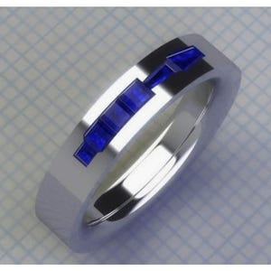 May include: A silver ring with a rectangular design of blue gemstones. The ring has a polished, reflective surface, and the blue stones are set in a linear pattern across the top of the band. The ring is shown on a grid background.