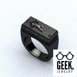 May include: A black ring featuring a rectangular top with a detailed relief of a figure. The ring has a thick band and a textured surface on the top. The Geek Jewelry logo is in the bottom right corner.