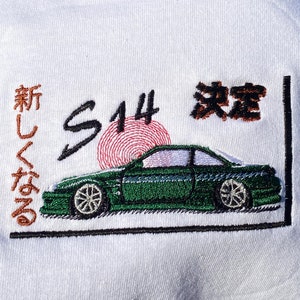 May include: A white t-shirt with an embroidered design featuring a green car, a red sun, and the text "S14" and Japanese characters. The text reads "新しくてなる".