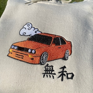 May include: A light gray hoodie with an embroidered orange car and a cloud of smoke above it. The text "無和" is embroidered below the car.