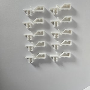 May include: A collection of ten white plastic clips arranged in rows. Each clip has a unique shape with a hook-like design on one end and a rectangular body. The clips are on a white surface, casting subtle shadows.