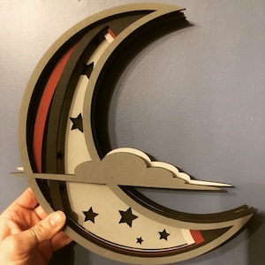 Puede incluir: Decoración de pared de luna creciente de madera en capas. El diseño presenta una combinación de colores gris, negro y rojo con estrellas y una nube recortadas. La luna se sostiene contra una pared azul.