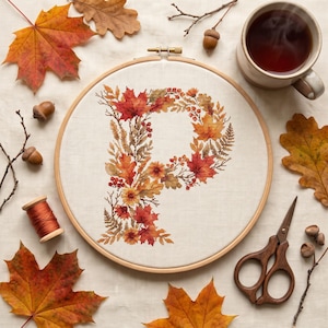 May include: A circular wooden embroidery hoop containing a cross-stitch design of the letter P, intricately formed by a collection of autumn leaves, berries, and small flowers in shades of orange, red, yellow, and brown on a light fabric background.