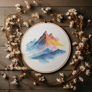 May include: A cross-stitch embroidery hoop with a colorful mountain landscape design. The mountains are a gradient of blue, purple, pink, orange, and yellow. The background is white.