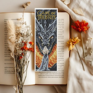 Puede incluir: Marcapáginas hecho a mano con un diseño pixelado que presenta un dragón y el texto "GAME OF THRONES". El marcapáginas está colocado sobre un libro abierto. También se ven flores secas.