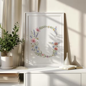 Puede incluir: Una impresión floral enmarcada con la letra "G" compuesta de flores coloridas. La obra de arte incluye flores rosas, azules y amarillas dispuestas en una fuente decorativa. La impresión se muestra en un mueble blanco con una planta y libros.