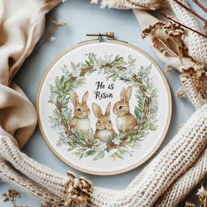 以下が含まれることがあります： 白い刺繍枠に、緑色の花の輪と3匹の茶色のウサギが描かれています。輪には白い花が飾られており、中央には「He is Risen」という文字が書かれています。