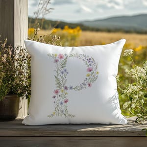 Può includere: Cuscino quadrato bianco con un design floreale a forma di "P". La lettera è composta da vari fiori nei toni del rosa, viola, giallo e verde. Il cuscino è appoggiato su una superficie di legno con uno sfondo naturale all'aperto.
